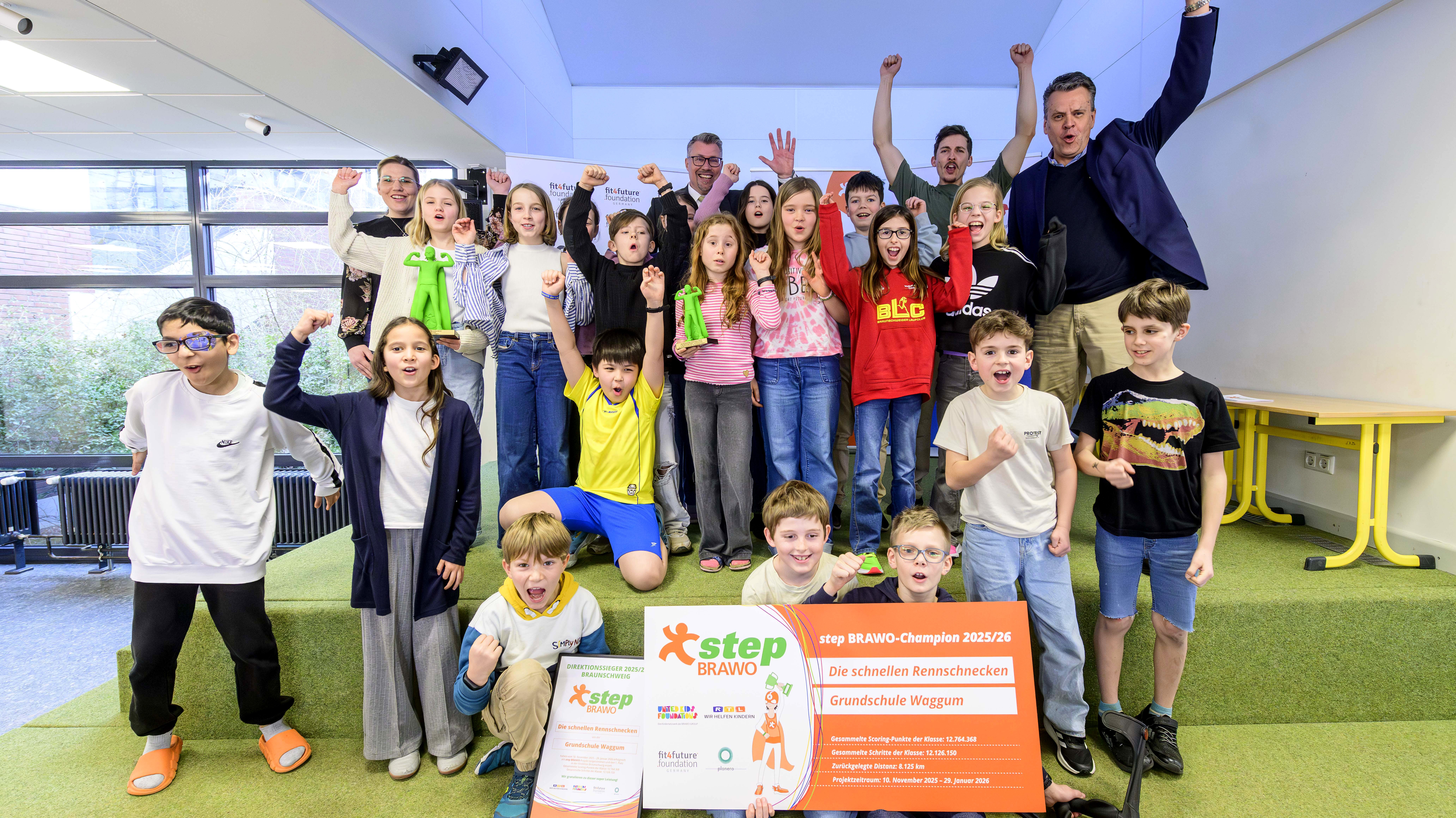 Eine Gruppe fröhlicher Kinder posiert zusammen mit einigen Erwachsenen in einem Raum auf einem Podest. Die Kinder tragen bunte Alltagskleidung und halten stolz ein großes orangenes Banner und eine Urkunde mit der Aufschrift "step BRAWO-Champion 2025/26". Einige Kinder haben ihre Fäuste in die Luft gereckt, offensichtlich in Feierstimmung.