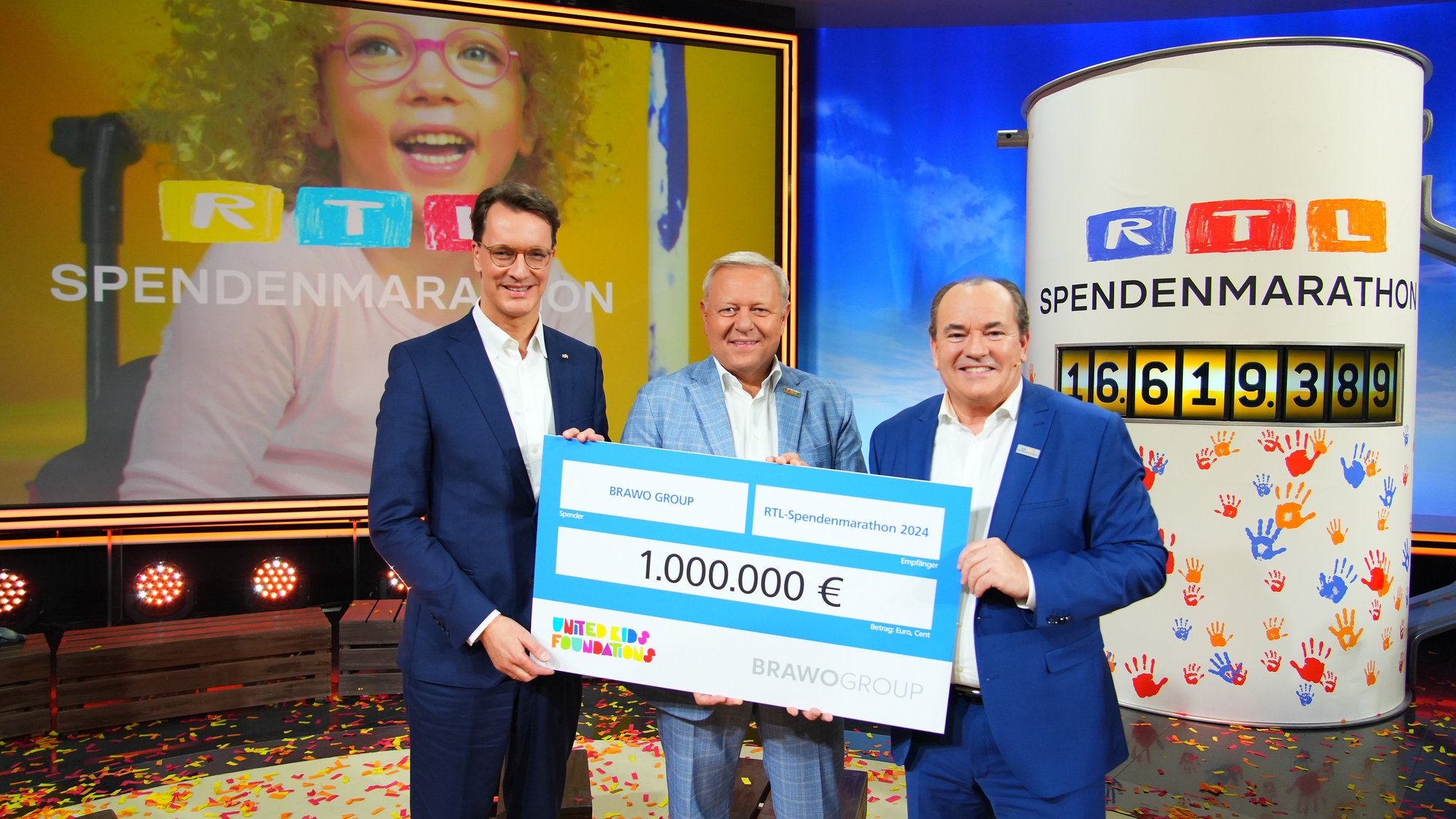 Drei Personen stehen nebeneinander und halten gemeinsam ein großes Schild, einem Spendenschein mit Wert von 1.000.000€, in der Hand. Sie stehen lächelnd vor einem bunten Hintergrund, in dem ein großen Bildschirm und einer Litfaßsäule. Auf dem Bildschirm ist ein lachendes Kind mit Brille zu sehen, davor ist das gelb-blau-rote-Logo von RTL zu sehen, darunter steht in weiß "Spendenmarathon". Das Logo ist ebenfalls auf der Litfaßsäule, darunter steht in schwarz "Spendenmarathon". Unter diesem Wort ist die Zahl "16619389" abgebildet. Wiederum darunter sind viele bunte Handabdrücke.