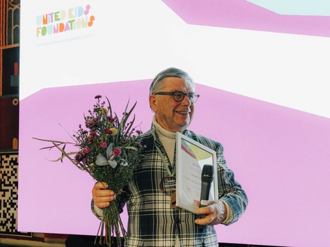 Ein älterer Herr steht vor einem großen Bildschirm, auf dem "United Kids Foundations" zusammen mit einem farbenfrohen Logo angezeigt wird. Er trägt ein kariertes Jackett, hält einen Blumenstrauß und eine Urkunde in der Hand. In der anderen Hand hält er ein Mikrofon. Im Hintergrund ist ein kunstvoll gestaltetes Wandbild zu sehen. Auf dem Bildschirm steht außerdem "20 Jahre" und "Das Kindernetzwerk der BRAVO GROUP".