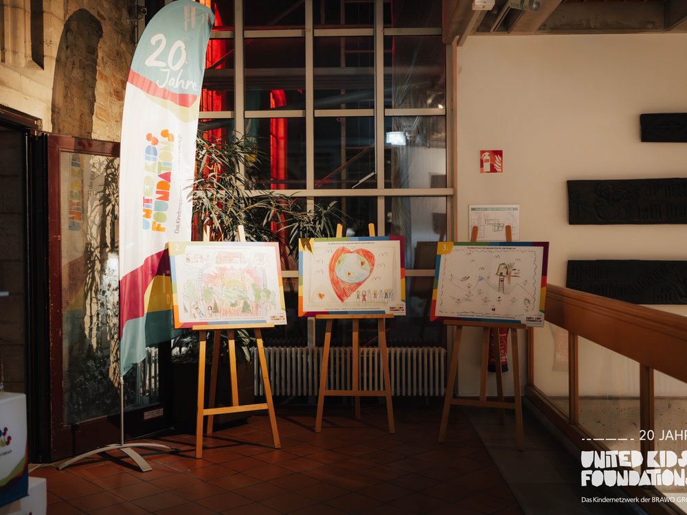Das Bild zeigt eine Ausstellung in einem gut beleuchteten Innenraum. Links steht ein Banner mit der Aufschrift "20 Jahre United Kids Foundations". Vor dem Banner befinden sich drei Staffeleien mit Kinderzeichnungen. Jede Zeichnung ist bunt gestaltet und zeigt verschiedene kreative Szenen. Im Hintergrund sind eine Pflanze und eine Fensterfront zu sehen. Rechts an der Wand hängt ein Feuerlöscherzeichen. Unten rechts im Bild steht "20 JAHRE UNITED KIDS FOUNDATIONS – Das Kindernetzwerk der BRAWO GROUP".