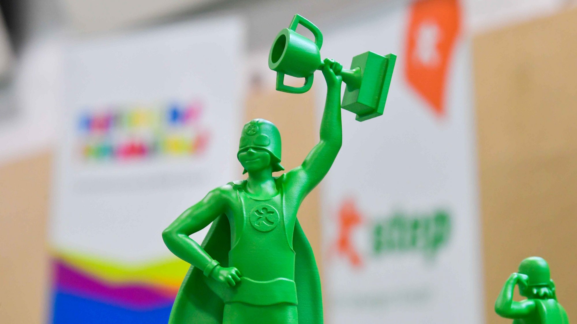 Das Bild zeigt eine grüne Superheldenfigur ab der Hüfte aufwärts. Die Figur streckt einen Arm nach oben und hält dort einen großen Pokal in der Hand. Die Figur trägt zusätzlich ein Cape und hat die die andere Hand in die Hüfte gesteckt.