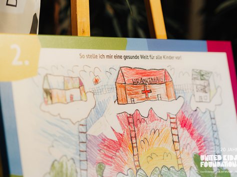 Eine kindliche Zeichnung, die eine Vorstellung von einer gesunden Welt darstellt. Auf dem Bild sieht man ein Krankenhaus auf einer Wolke mit einem roten Kreuz und der Aufschrift "Krankenhaus". Links sind bunte Gebäude ebenfalls auf Wolken zu sehen. Im Hintergrund gibt es eine bunte Landschaft mit Treppen, die zu den Wolken führen, und einem Sonnenaufgang. Oben steht: "So stelle ich mir eine gesunde Welt für alle Kinder vor!" Unten rechts ist das Logo der "United Kids Foundations" mit dem Hinweis auf das 20-jährige Jubiläum.