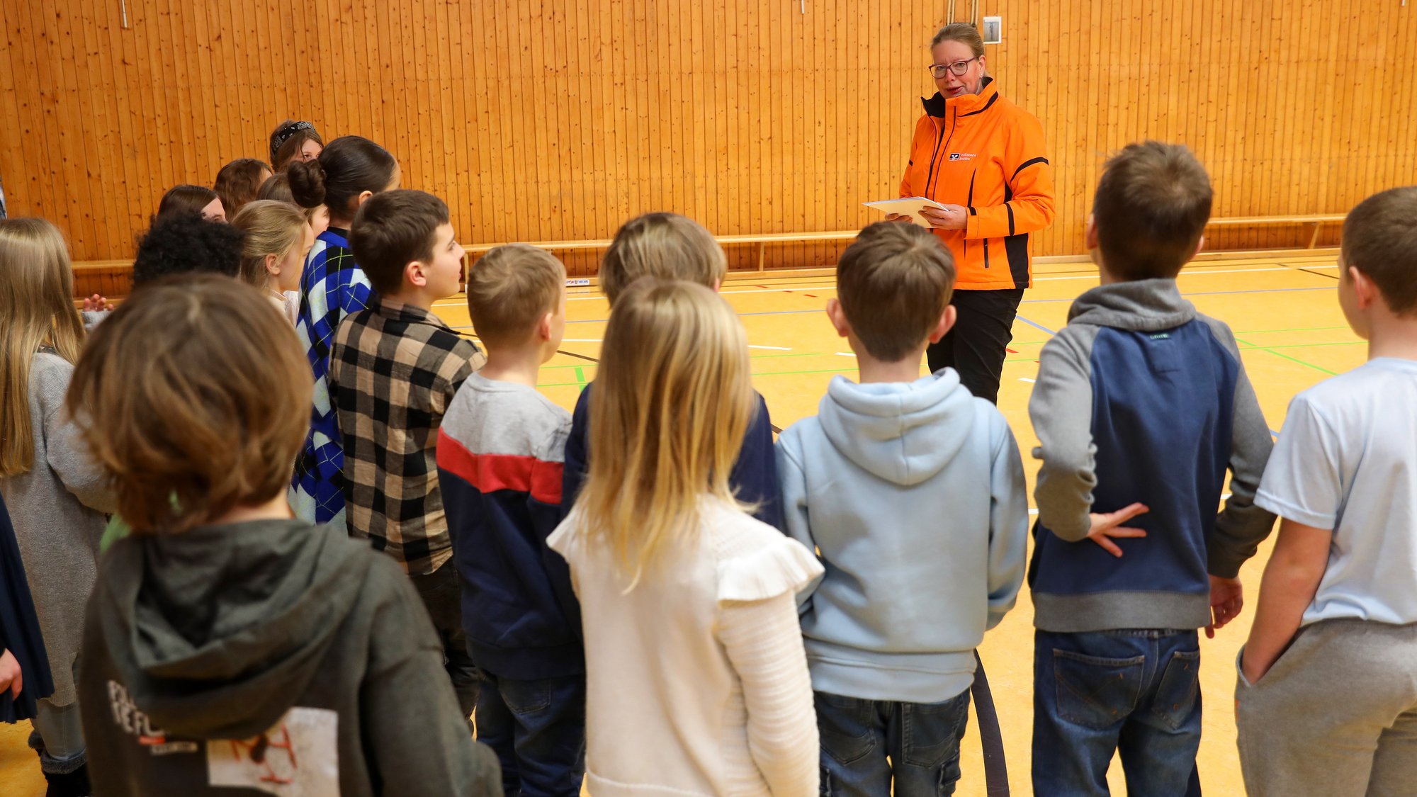 Auf dem Bild steht eine Frau in orangefarbener Jacke vor einer Gruppe Kinder, die nur von hinten zu sehen sind. Sie stehen in einer Sporthalle.