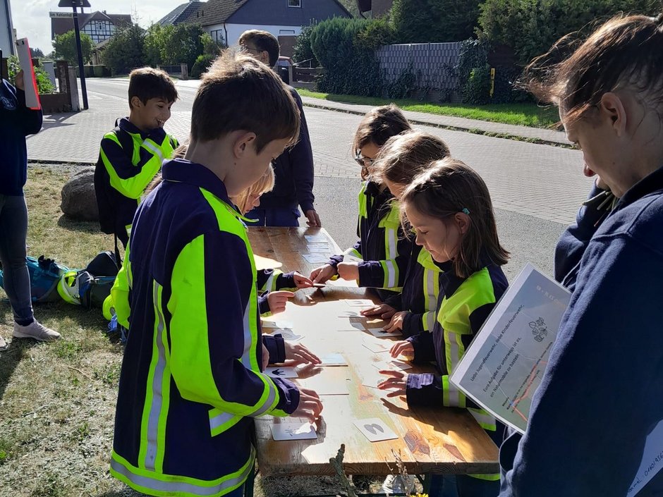 Eine Gruppe von Kindern stehen an einem Holztisch. Vor ihnen liegen verschiedene Kärtchen. Die Kinder tragen eine Feuerwehrjacke mit auffälligen gelben Streifen.