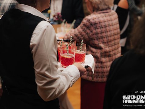 Eine Servicekraft trägt ein Tablett mit mehreren roten Getränken in Gläsern, die grün-weiß gestreifte Strohhalme haben. Im Hintergrund sind mehrere Personen bei einer Veranstaltung zu sehen. Unten rechts im Bild befindet sich ein Schriftzug mit dem Logo der "United Kids Foundations" und einem Hinweis auf das 20-jährige Jubiläum.