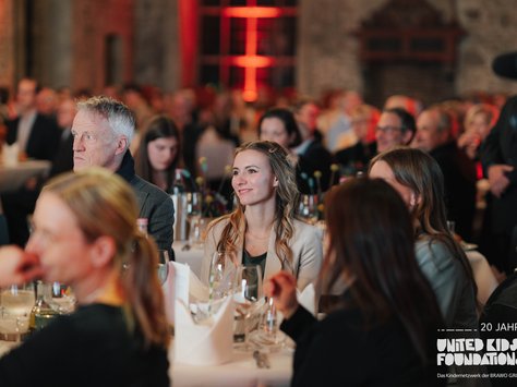 Ein festlich dekorierter Raum mit zahlreichen Menschen, die an Tischgruppen sitzen. Viele von ihnen schauen aufmerksam nach vorne. Eine Frau in der Mitte trägt ein helles Jackett und lächelt. Auf den Tischen stehen Weingläser, Flaschen und Blumenarrangements. In der unteren rechten Ecke ist das Logo von "United Kids Foundations" mit dem Hinweis auf das 20-jährige Jubiläum abgebildet.