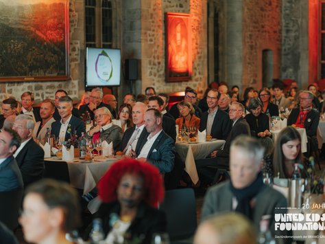 In einem festlich beleuchteten Raum sitzen viele Menschen an runden Tischen, offenbar bei einer Veranstaltung. Die Wände sind aus Stein, und Gemälde hängen daran. Im Hintergrund ist ein Bildschirm mit einem Logo zu sehen. Die Gäste scheinen aufmerksam einer Präsentation zu folgen. Auf der rechten unteren Ecke steht das Logo von "United Kids Foundations" mit dem Hinweis auf das 20-jährige Jubiläum.
