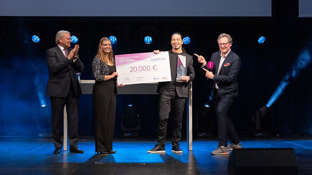 Vier Personen stehen lächelnd auf einer Bühne. Die beiden Personen in der Mitte halten einen großen Scheck auf dem 20.000€ steht hoch.