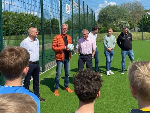 Im Vordergrund ist eine Gruppe Kinder zu sehen, die mit dem Rücken zur Kamera stehen. Sie schauen auf, in einer Reihe stehende, Erwachsene, von denen einer von ihnen einen Fußball in der Hand hält und etwas erzählt. Alle stehen auf einer Grünfläche, mit einem hohen Zaun im Hintergrund.