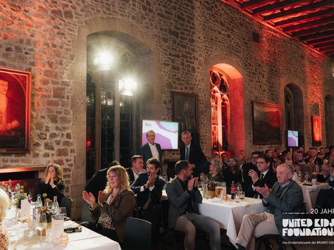 Das Bild zeigt eine festliche Veranstaltung in einem historischen Raum mit Steinwänden und großen Fenstern. Zahlreiche Personen sitzen an langen, weiß gedeckten Tischen. Einige Personen klatschen Beifall. Zwei Personen stehen in der Nähe eines Monitors, auf dem ein Präsentationsbild zu sehen ist. Der Raum ist stimmungsvoll in Rot- und Orangetönen beleuchtet. An den Wänden hängen mehrere Gemälde. Im unteren rechten Bereich des Bildes befindet sich ein Logo mit der Aufschrift "20 JAHRE UNITED KIDS FOUNDATIONS, Das Kindernetzwerk der BRAWO GROUP".