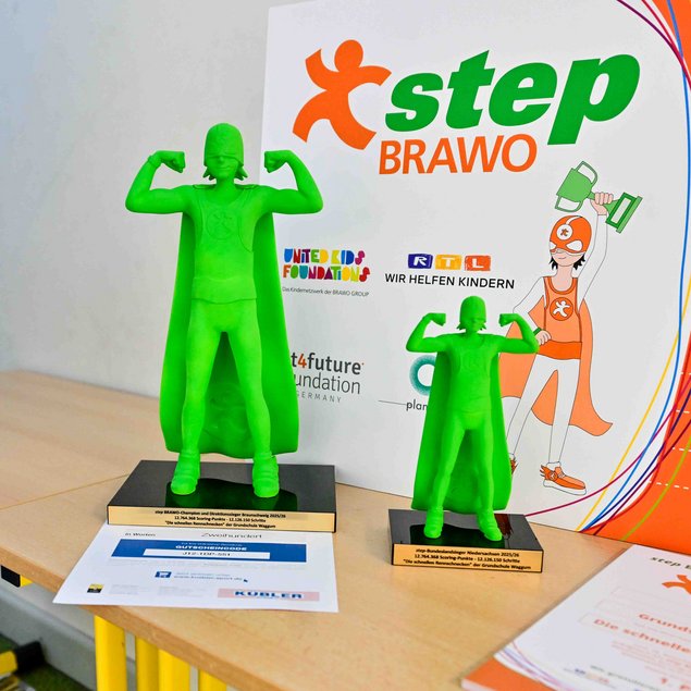Auf einem Tisch stehen zwei grüne Trophäen in Form eines Superhelden mit Umhang, der die Arme in einer Siegerpose hebt. Hinter ihnen befindet sich ein farbenfrohes Poster mit der Aufschrift "step BRAVO", gefolgt von Logos und weiteren Informationen. Auf dem Tisch liegt auch ein Zertifikat. Das Thema scheint sich um eine Auszeichnung oder ein Projekt im Zusammenhang mit Kindern und Gesundheitsförderung zu drehen.