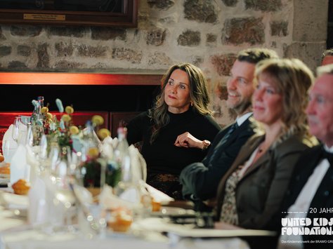 Das Bild zeigt eine festliche Tafel in einem Raum mit Steinwänden. Mehrere Menschen sitzen an einem langen, gedeckten Tisch, der mit Wasserflaschen, Gläsern und Blumen dekoriert ist. Eine Frau im schwarzen Oberteil sitzt aufmerksam und blickt leicht zur Seite, während andere Anwesende lächeln und ebenfalls interessiert wirken. Im Vordergrund sind diverse Teller und Servietten zu sehen. In der unteren Ecke des Bildes befindet sich das Logo "United Kids Foundations" mit einem Hinweis auf 20 Jahre Jubiläum.