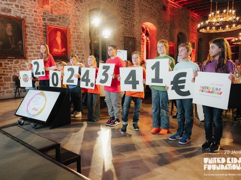 In einem Saal stehen sieben Kinder nebeneinander, die große Plakate mit Ziffern und einem Eurozeichen hochhalten, die zusammen einen Geldbetrag von 2.024.341 Euro ergeben. Die Kinder tragen bunte Kleidung. Im Hintergrund sind Menschen an Tischen und Gemälde an den Steinwänden zu sehen. Unten rechts wird auf das 20-jährige Jubiläum der "United Kids Foundations" hingewiesen. Der Raum ist warm beleuchtet und hat große Kronleuchter an der Decke.