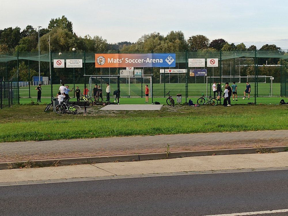 Umzäunte Fußballplätze, auf denen mehrere Gruppen von Kindern stehen. Auf einem Plakat am Zaun steht "Matts Soccer Arena".