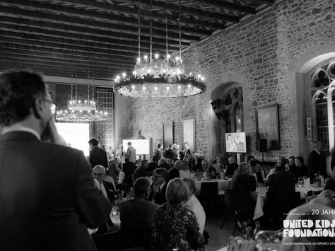 Ein großer, gut beleuchteter Raum mit einer hohen Decke und mehreren Kronleuchtern. Viele Menschen in formeller Kleidung sitzen an runden Tischen, die für ein Event gedeckt sind. Die Wände sind aus Ziegelsteinen und schmücken sich mit großen Fenstern und Bildern. Im Vordergrund sind Personen zu sehen, die miteinander sprechen, während im Hintergrund eine Präsentation auf Bildschirmen zu erkennen ist. Die Atmosphäre wirkt geschäftig und festlich. In der Ecke befindet sich das Logo "United Kids Foundations" sowie der Hinweis auf das 20-jährige Jubiläum.
