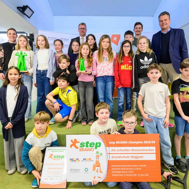 Eine Gruppe von Kindern und Erwachsenen posiert fröhlich für ein Foto. Die Kinder stehen und sitzen auf einer grünen Stufenfläche. Zwei Kinder halten grüne Trophäen. Im Vordergrund präsentiert ein Kind ein großes Schild, das auf einen Wettbewerb hinweist. Die Erwachsenen stehen hinter den Kindern und lächeln. Im Hintergrund sind Fenster und weitere dekorative Elemente sowie Banner zu sehen.