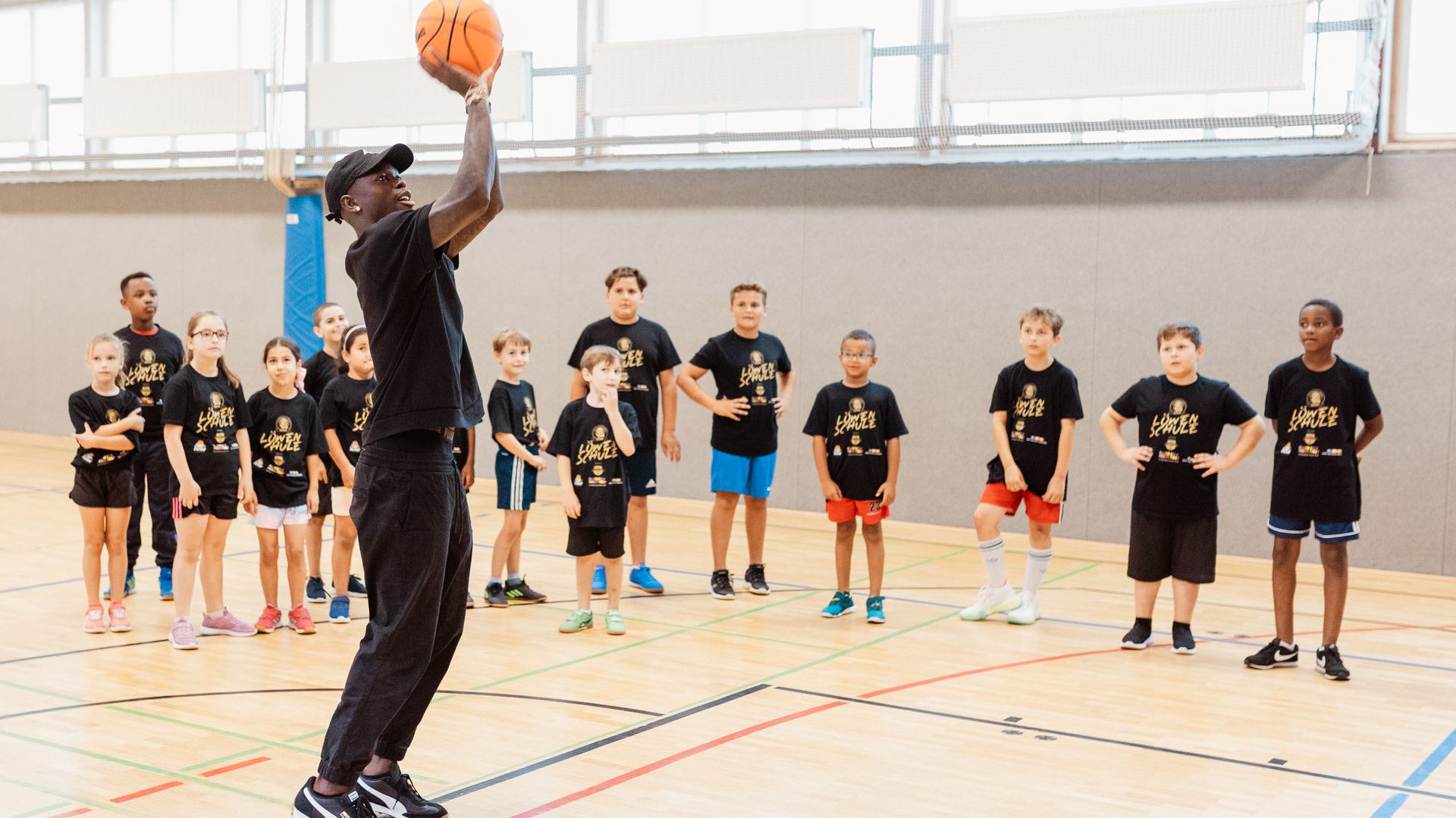 Dennis Schröder wirft einen Basketball in einer Turnhalle. Eine Gruppe von Kindern in schwarzen T-Shirts beobachtet aufmerksam. Die Halle ist mit einem Holzboden und großen Fenstern ausgestattet.