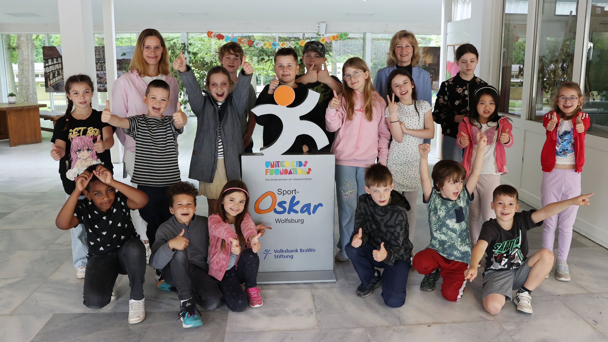 Gruppe von Kindern und zwei Erwachsenen posieren fröhlich um ein Schild mit der Aufschrift 'Sport Oskar Wolfsburg' in einem hellen Innenraum.
