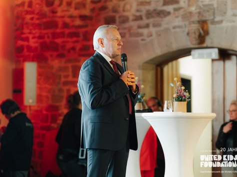 Ein älterer Mann steht in einem Raum mit Ziegelwänden und spricht in ein Mikrofon. Er trägt einen Anzug und eine Krawatte. Im Hintergrund sind einige Leute und ein hoher Tisch mit Blumendekoration zu sehen. Auf dem Bild steht "20 Jahre United Kids Foundations".