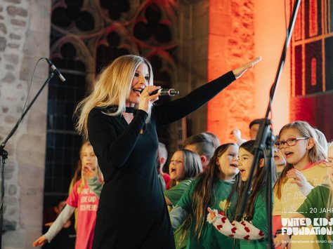 Eine blonde Frau singt mit einem Mikrofon auf einer Bühne. Um sie herum stehen mehrere Kinder, die ebenfalls singen. Die Szenerie ist festlich beleuchtet, mit warmen Farben und einer gemauerten Wand im Hintergrund. Eine Aufschrift am rechten unteren Rand des Bildes weist auf den 20. Jahrestag der United Kids Foundations hin.