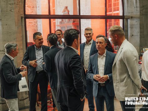 Eine Gruppe von Männern in Anzügen steht in einem festlichen Raum und unterhält sich. Die Stimmung wirkt gesellig und konzentriert. Im Hintergrund sind große Fenster zu sehen, die einen hellen Schein in den Raum werfen. Einige der Männer halten Getränke in den Händen. Auf dem Bild ist ein Logo mit der Aufschrift "20 Jahre United Kids Foundations" zu sehen.