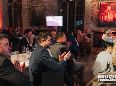 Eine Gruppe von Menschen sitzt an runden Tischen in einem historischen Raum mit Steinmauern und bunten Lichteffekten. Die Gäste tragen formelle Kleidung und klatschen, während sie auf eine Präsentation oder Rede fokussiert sind. Ein Gemälde hängt an der Wand und ein Bildschirm mit einer grafischen Darstellung ist im Hintergrund zu sehen. In der Ecke des Bildes befindet sich ein Logo von "United Kids Foundations" mit einem Hinweis auf das 20-jährige Jubiläum.