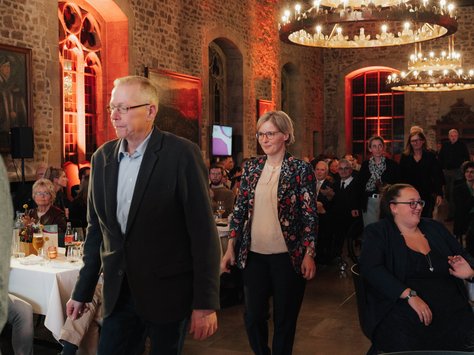 In einem festlich beleuchteten Saal mit Steinmauern und großen Kronleuchtern gehen mehrere Personen an einem gedeckten Tisch entlang. Einige Gäste sitzen an Tischen und unterhalten sich. Im Hintergrund sind dekorative Gemälde und Fenster zu sehen. Ein Banner zeigt das Logo von "United Kids Foundations".