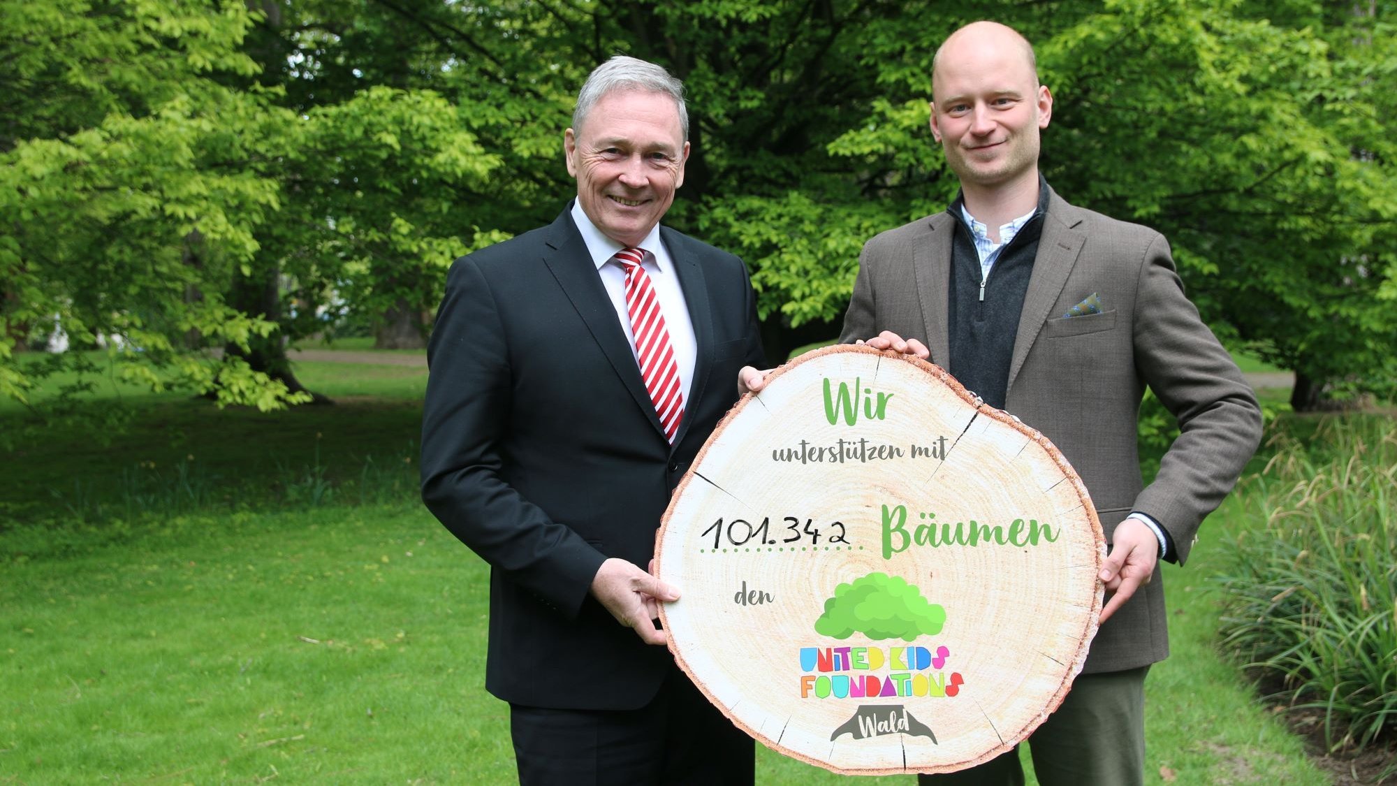 Zwei Personen im Anzug stehen nebeneinander auf einer grünen Wiese vor einigen Bäumen. Sie halten ein abgesägten Baumstamm mit der Aufschrift "Wir unterstützen mit 101.342 Bäumen den United Kids Foundations Wald".