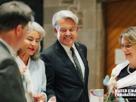 Eine Gruppe von Personen steht zusammen und unterhält sich in einem formellen Rahmen. Alle wirken gut gekleidet und lächeln. Im Vordergrund ist ein Blumenarrangement zu sehen. In der unteren rechten Ecke ist das Logo "United Kids Foundations" sichtbar.