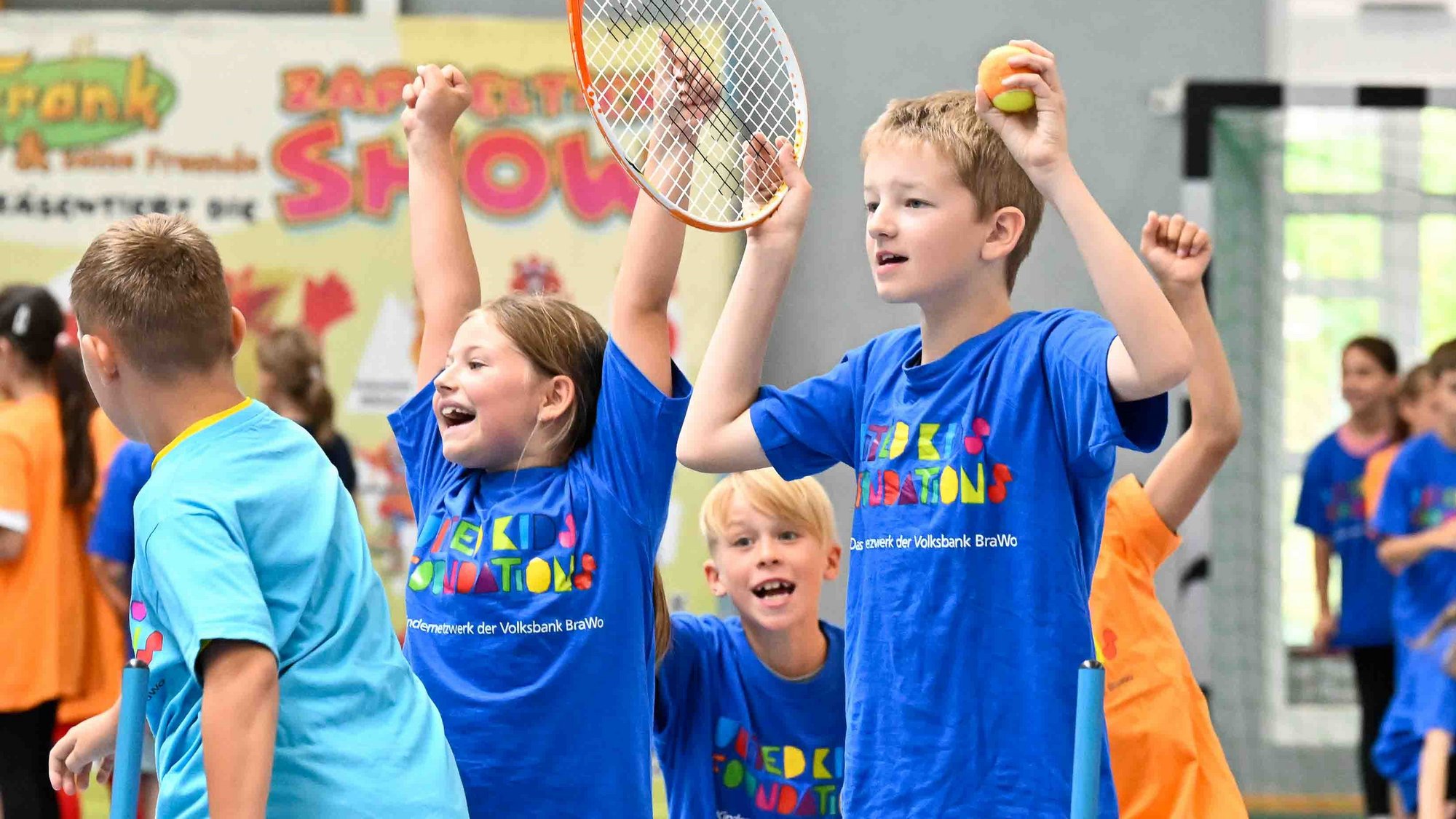 Die Schülerinnen und Schüler der Grundschule Jembke hatten viel Spaß beim Sport-Oskar. Credit: Volksbank BRAWO/Isabell Massel Sich freuende Kinder in bunten Tshirts in einer Sporthalle. Ein Kind hält einen Tennisball in der linken und einen Tennisschläger in der rechten Hand. Den gleichen Tennisschläger hält auch ein weiteres Kind neben ihm fest.