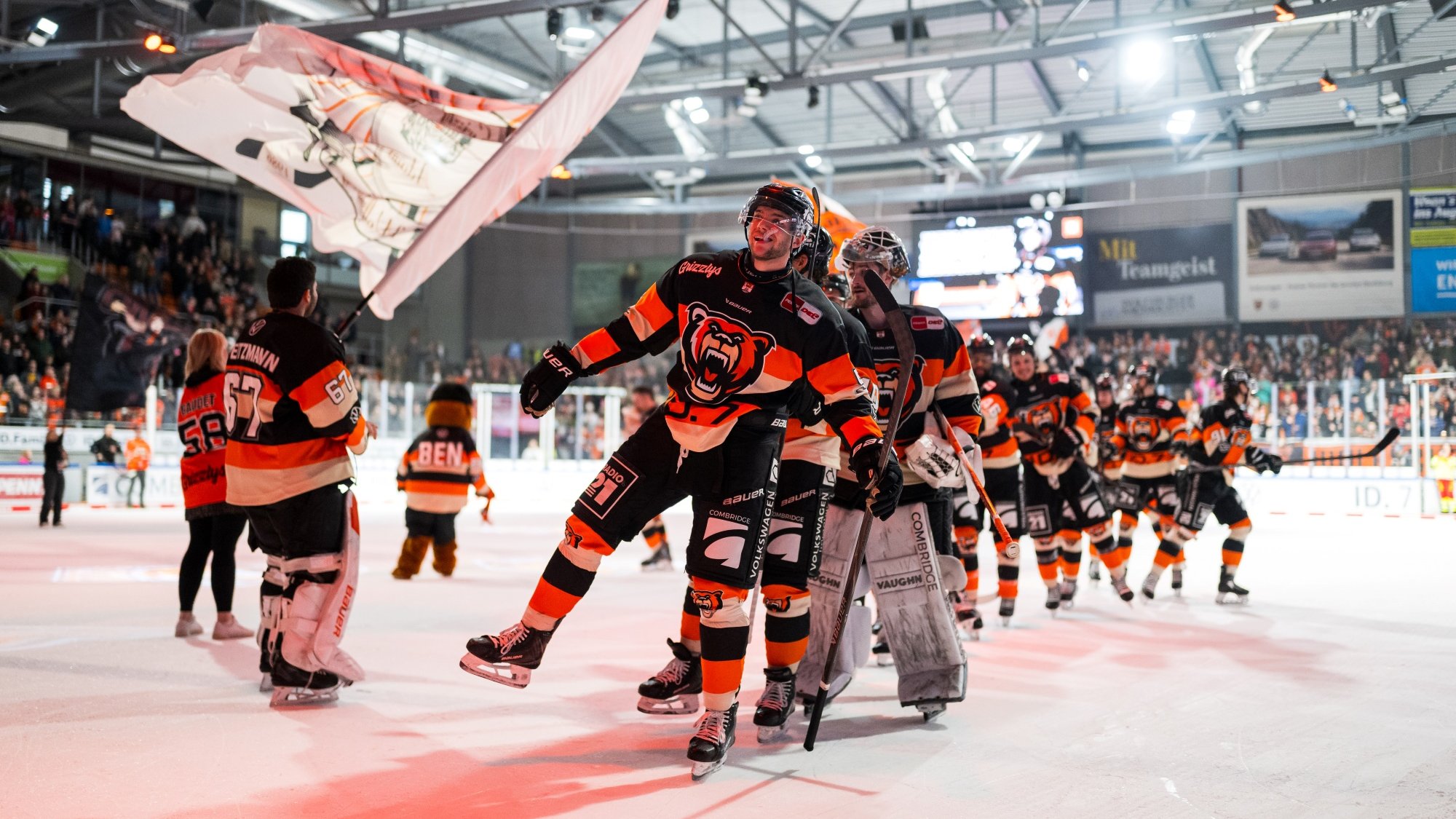 Eine Eishockeymannschaft in schwarz-orangen Trikots feiert auf dem Eis. Die Spieler haben Eishockeyausrüstung an und scheinen gerade ein Spiel gewonnen zu haben. Im Vordergrund schwenkt eine Person eine große Fahne. Im Hintergrund sind jubelnde Fans in einer vollbesetzten Eishalle zu sehen. Die Atmosphäre ist fröhlich und triumphierend.