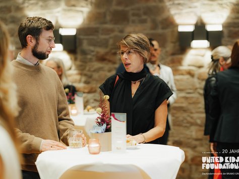 Im Bild befinden sich zwei Personen, die sich an einem festlich gedeckten Stehtisch unterhalten. Der Mann trägt einen beigefarbenen Pullover und hält ein Glas in der Hand. Die Frau trägt eine schwarze Bluse und hat kurze Haare. Auf dem Tisch stehen Kerzen und florale Dekorationen. Im Hintergrund sieht man eine Steinwand und einige weitere Gäste. Rechts unten ist das Logo „United Kids Foundations - 20 Jahre“ zu sehen.