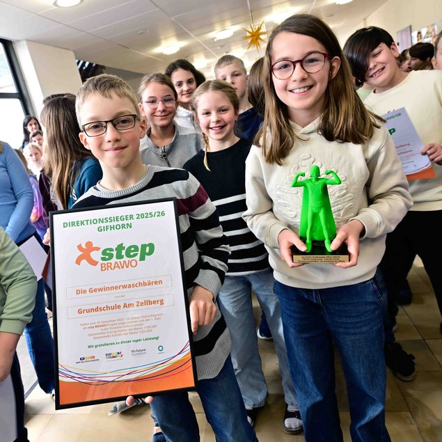 Eine Gruppe von Schülern steht in einem Flur. Ein Kind hält ein Zertifikat mit der Aufschrift „Direktionssieger 2025/26 Gifhorn“ und „step BRAWO“. Ein anderes Kind hält eine grüne Trophäe in Form einer Figur. Die Schüler lächeln und wirken stolz. Im Hintergrund sind weitere Schüler zu sehen.