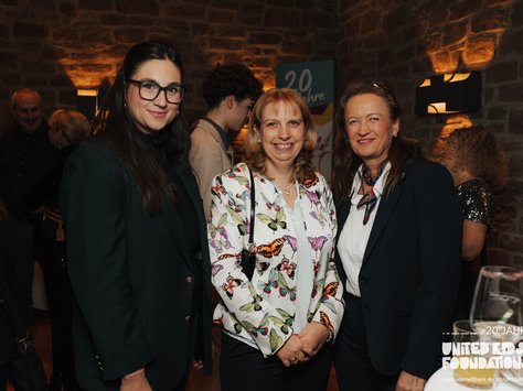 Drei Personen stehen zusammen und lächeln in die Kamera. Die linke Person trägt eine Brille und einen dunklen Blazer, die mittlere Person trägt eine Jacke mit buntem Schmetterlingsmuster, und die rechte Person trägt einen dunklen Blazer mit einem Tuch um den Hals. Im Hintergrund sind weitere Personen in einem stimmungsvoll beleuchteten Raum zu sehen. Rechts unten im Bild befindet sich ein Logo mit der Aufschrift "20 Jahre United Kids Foundations".