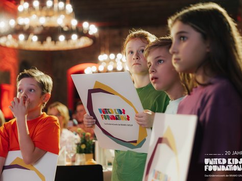 Vier Kinder stehen nebeneinander in einem festlich beleuchteten Raum und halten Schilder mit dem Logo der "United Kids Foundations". Die Kinder schauen gespannt in dieselbe Richtung. Im Hintergrund sind unscharf einige Gäste und ein großer Kronleuchter zu sehen. Das Bild vermittelt eine Atmosphäre der Aufmerksamkeit und Neugierde.