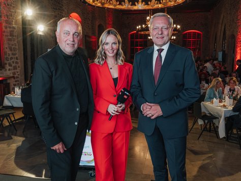 In einem eleganten Raum mit Steinwänden und stilvoller Beleuchtung stehen drei Menschen vor der Kamera. Links steht ein Mann in einem schwarzen Anzug, in der Mitte eine Frau in einem leuchtend roten Anzug mit einem Mikrofon, und rechts ein Mann in einem dunkelblauen Anzug mit roter Krawatte. Im Hintergrund sind Tische und Menschen zu sehen, die in einer festlichen Atmosphäre zusammenkommen. Unten rechts im Bild ist ein Logo mit der Aufschrift "20 Jahre United Kids Foundations" sichtbar.