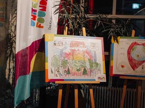 Ein farbenfrohes Kinderbild ist auf einer Staffelei ausgestellt. Daneben steht ein Banner mit der Aufschrift "United Kids Foundations". Das Bild zeigt eine bunte Landschaft mit Bäumen und Häusern. Im Hintergrund sind Pflanzen und ein großes Fenster zu sehen. Unten rechts steht das Logo der "United Kids Foundations" mit "20 Jahre" darüber. Der Raum ist warm beleuchtet.