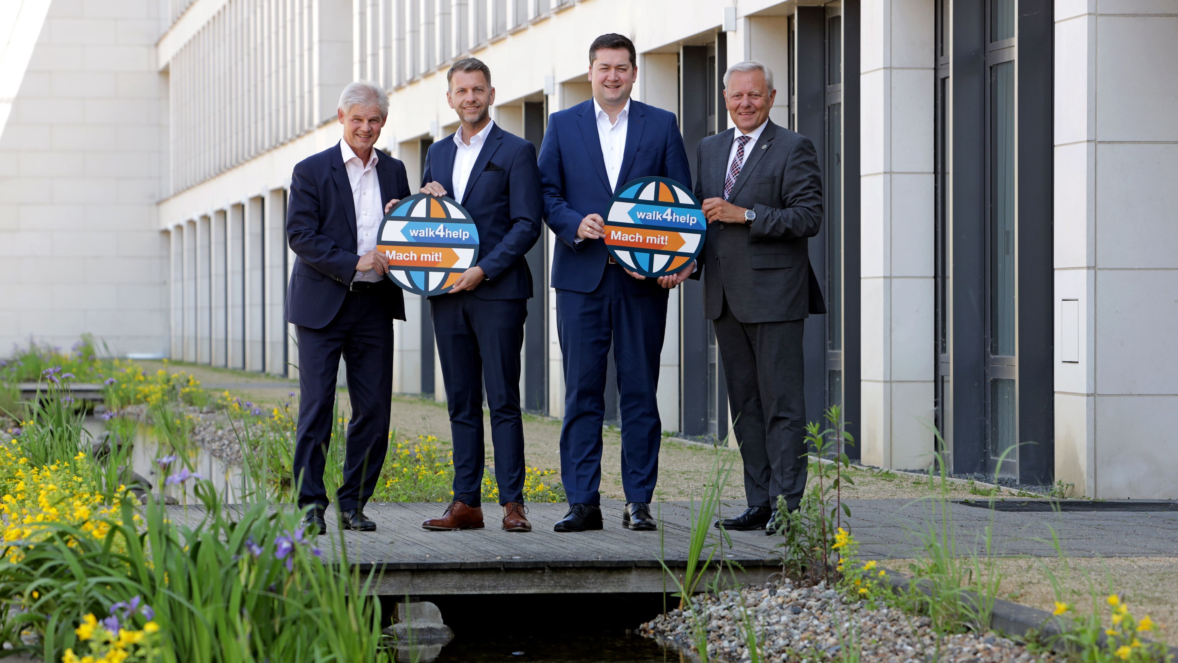 Die Oberbürgermeister Frank Klingebiel, Dennis Weilmann und Dr. Thorsten Kornblumen stehen in einer Reihe neben Jürgen Brinkmann auf einer kleinen Holzbrücke. Darunter ist ein Bach, im Hintergrund ein Gebäude zu sehen.