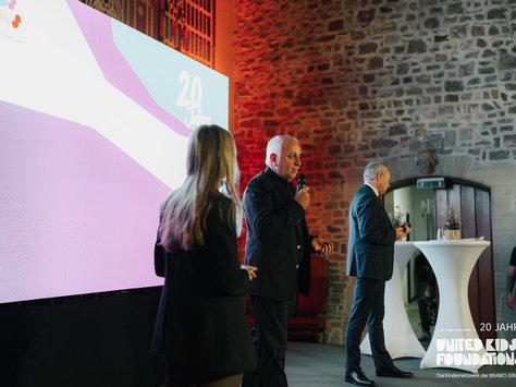 Auf dem Bild befindet sich eine Veranstaltung zum 20-jährigen Jubiläum der United Kids Foundations. Im Vordergrund steht ein Mann in einem dunklen Anzug, der in ein Mikrofon spricht. Eine Frau steht ebenfalls auf der Bühne, mit dem Rücken zur Kamera. Hinter ihnen ist eine große Leinwand mit der Aufschrift "20 Jahre United Kids Foundations" zu sehen. Der Raum hat steinerne Wände und einen eleganten, historischen Charakter. Ein weiterer Mann im Anzug läuft an einem Stehtisch vorbei, im Hintergrund sind weitere Platten an der Wand sichtbar.
